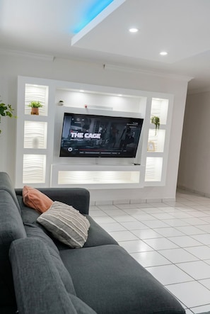 A modern living room with a flat-screen TV mounted on a white wall, a grey sofa with a cushion, and a tiled floor.