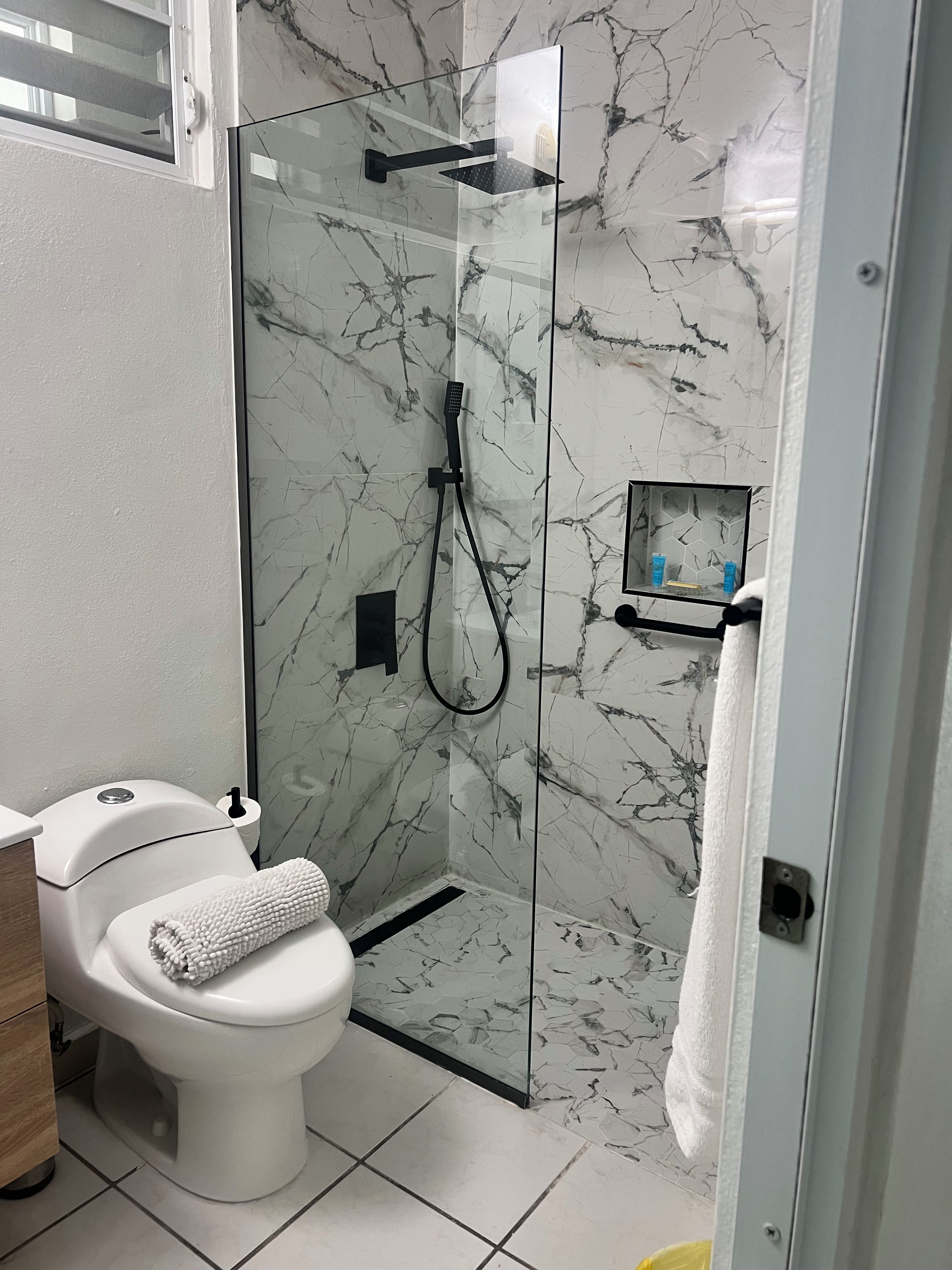 A modern bathroom with a glass-enclosed shower, a toilet, and a small shelf with toiletries.