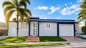 Une maison moderne à deux étages, avec une façade bleue et blanche, une porte d’entrée, un garage et une pelouse bien entretenue agrémentée de palmiers.