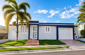 A modern two-story house with a blue and white exterior, a front door, a garage, and a well-maintained lawn with palm trees.
