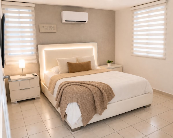A modern bedroom with a large bed, bedside tables, an air conditioner, and a motivational quote on the wall.