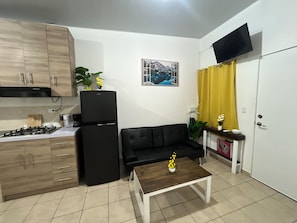 Ein kompakter Wohnraum mit Küchenzeile, einem schwarzen Sofa, einem Couchtisch und einem an der Wand befestigten Fernseher.