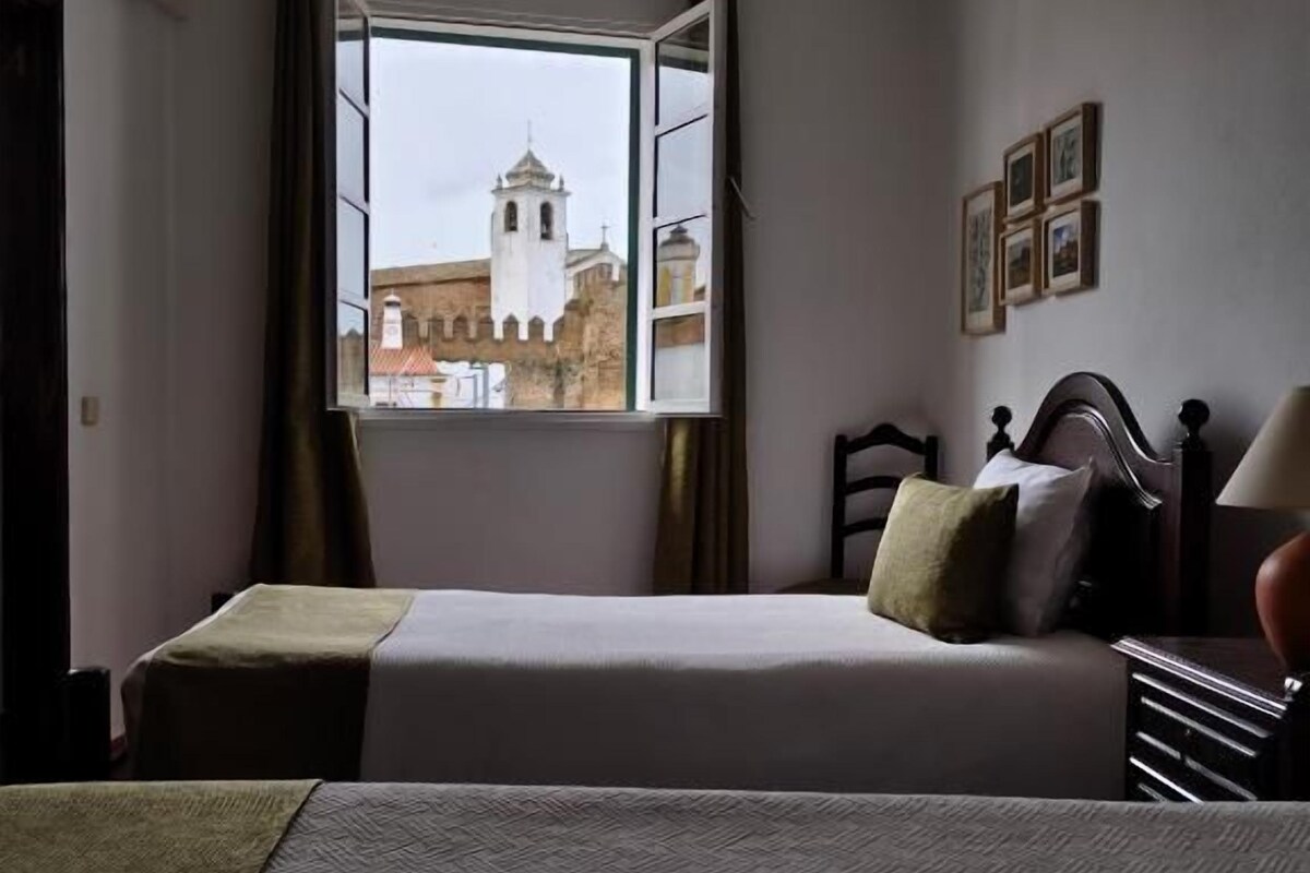 Een hotelkamer met een bed, een raam met gordijnen, een nachtkastje en een wandlamp.