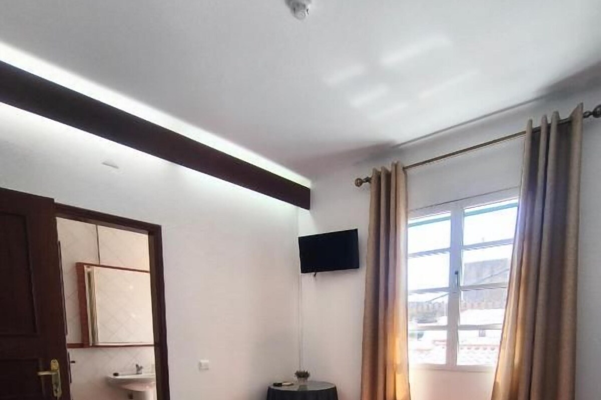 Een hotelkamer met een bed, een flatscreen televisie aan de muur, een raam met gordijnen en een deur naar een andere kamer.