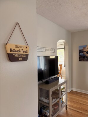 A living room with a television on a stand, a bookshelf, and a wall sign indicating the location as Deschutes National Forest, Three Creek Lake. - The Blackberry Cottage 2-bedroom house in beautiful Bend for relaxing stays! (Bend)