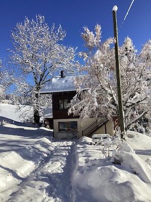A snow-covered house with a visible staircase leading to the entrance, a clear blue sky, and surrounding snow-laden trees. - New Listing (Autrans-Méaudre en Vercors)