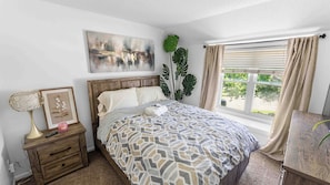 A bedroom with a bed, bedside table, lamp, artwork, and a window with curtains. - Spacious 5-bedroom house in serene Kansas City (Kansas City)