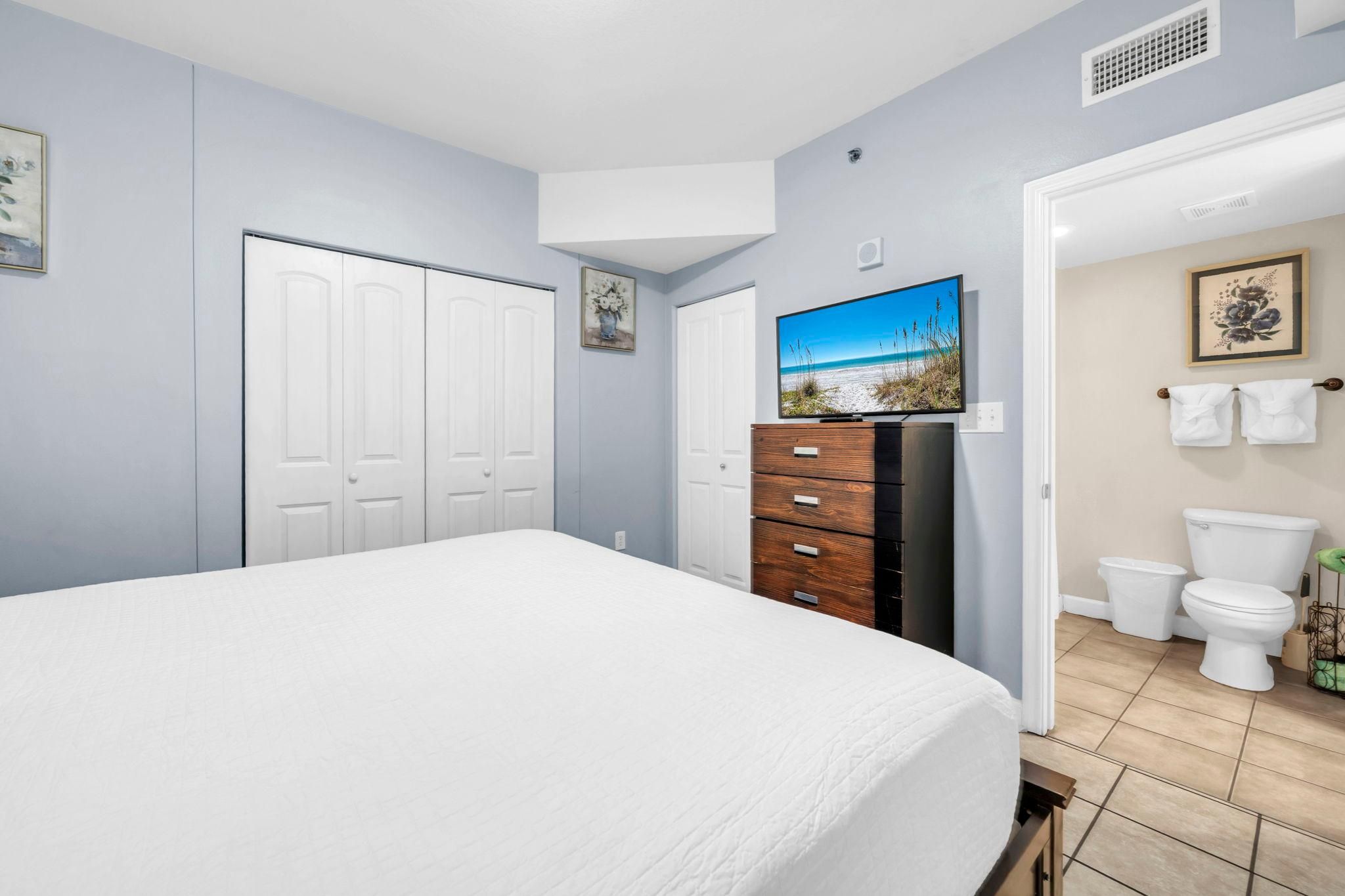 A bedroom with a bed, a dresser, a television, and a bathroom with a toilet and sink.