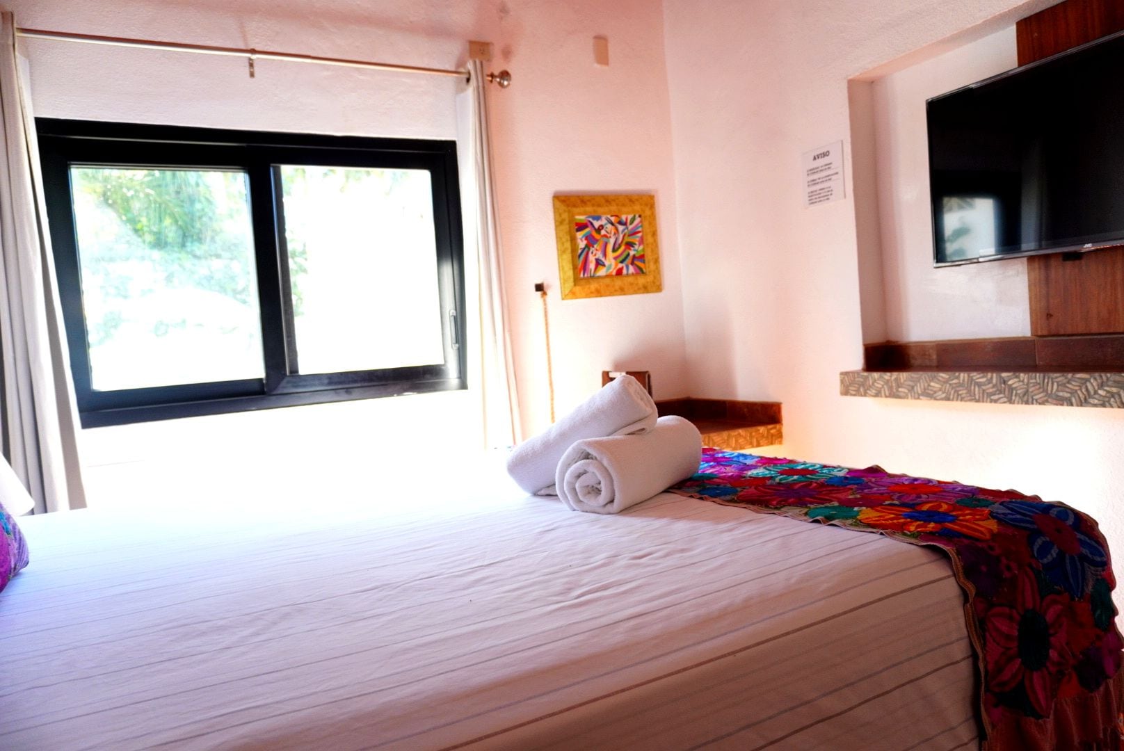 A hotel room with a bed, a television, a window, and a painting on the wall.