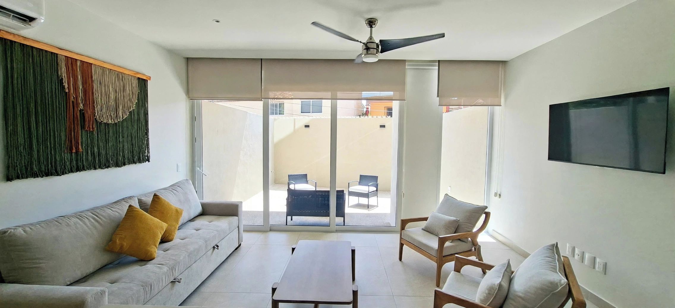 A modern living room with a sofa, a coffee table, and two chairs. There is a ceiling fan and a TV mounted on the wall.
