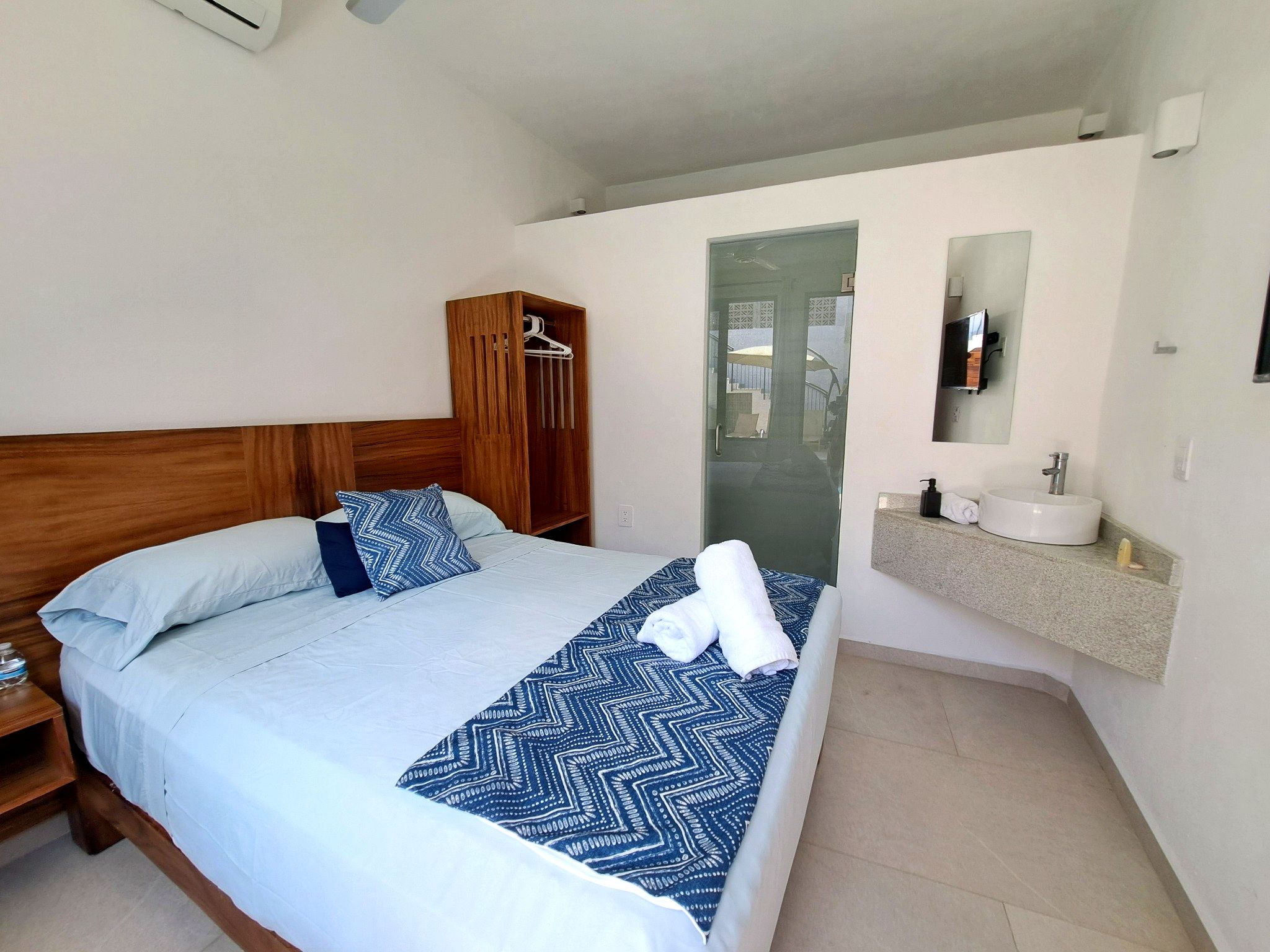 Habitació d'hotel amb un llit, un armari de fusta, un bany amb un rentapeus i un mirall, i una vista a través d'una porta de vidre.