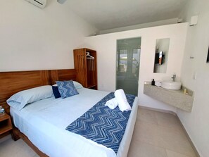 Habitación de hotel con cama, armario de madera, baño con lavamanos y espejo, y vistas a través de una puerta de cristal.