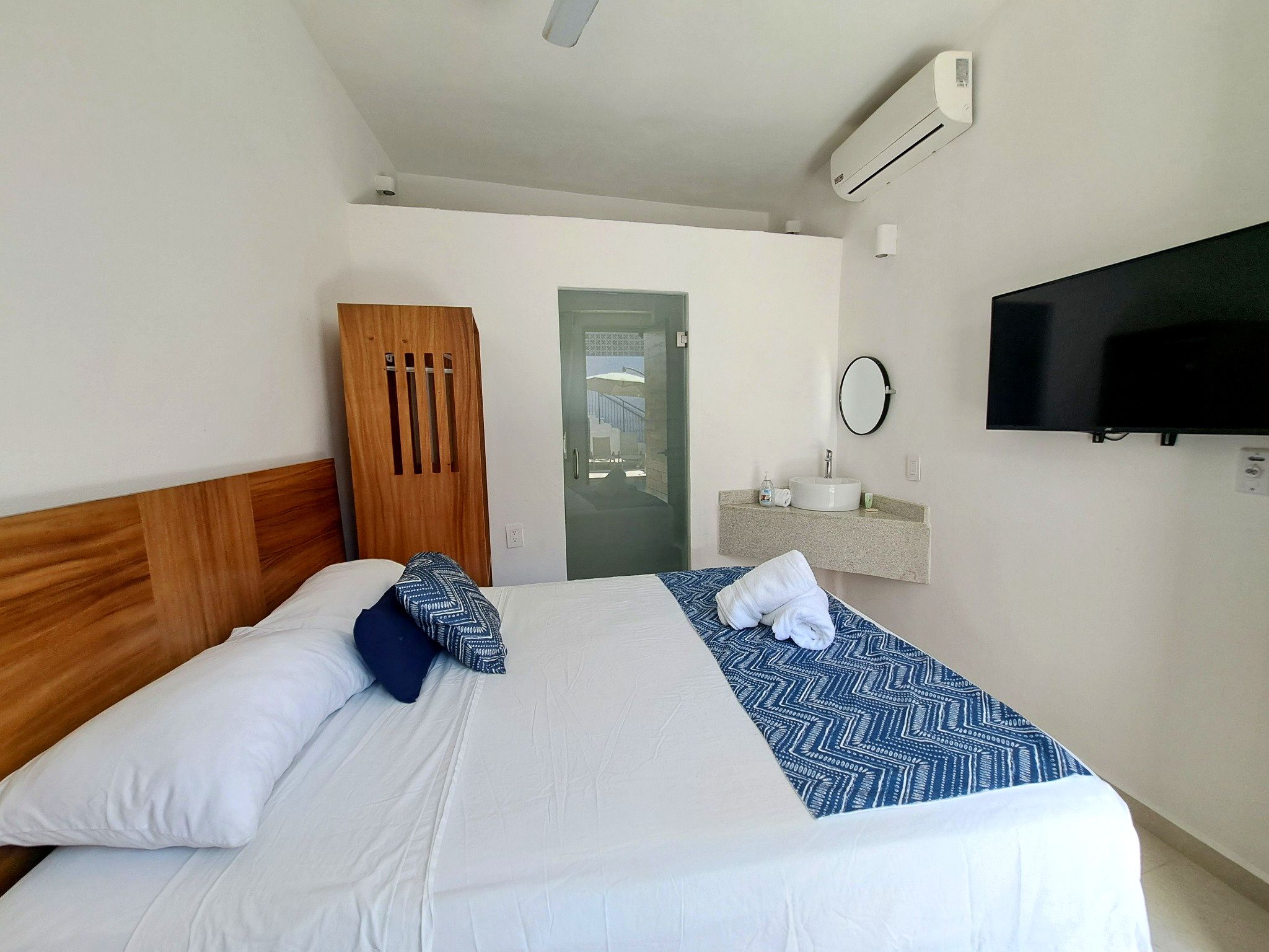 A hotel room with a bed, a wooden wardrobe, a bathroom with a sink and mirror, and a TV mounted on the wall.