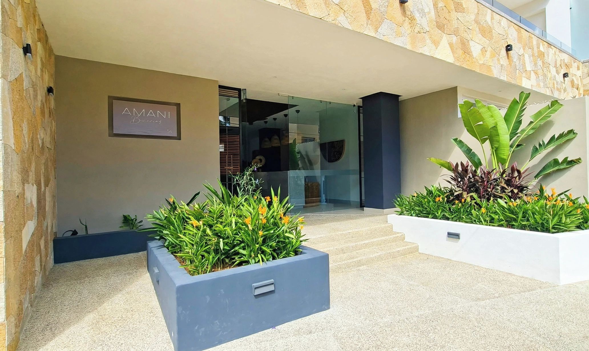 A modern building entrance with a stone facade, a sign for 'AMANI Brunches,' and two planters with greenery.