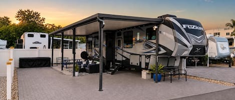 A covered RV parking area with a modern design, including a covered seating area with chairs and a table, and a large RV with the brand name 'FUXION' visible.