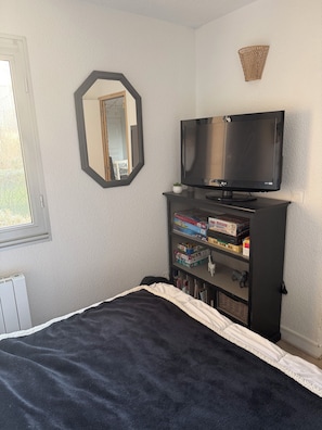 Een slaapkamer met een bed, een televisie op een standaard, een plank met gezelschapsspellen, en een raam.