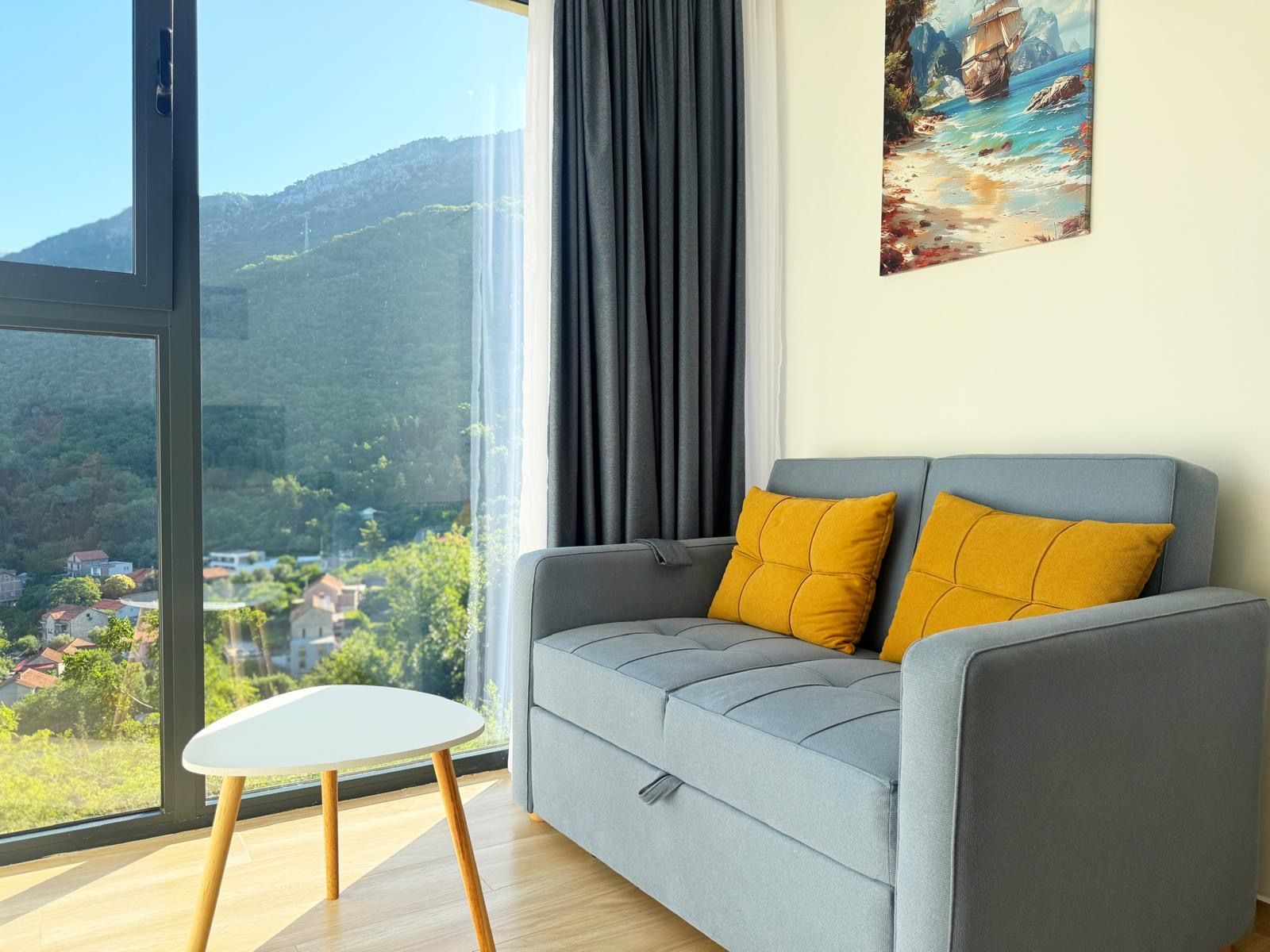 A modern living room with a grey sofa, two mustard pillows, a white side table, and a large window offering a view of a mountainous landscape.