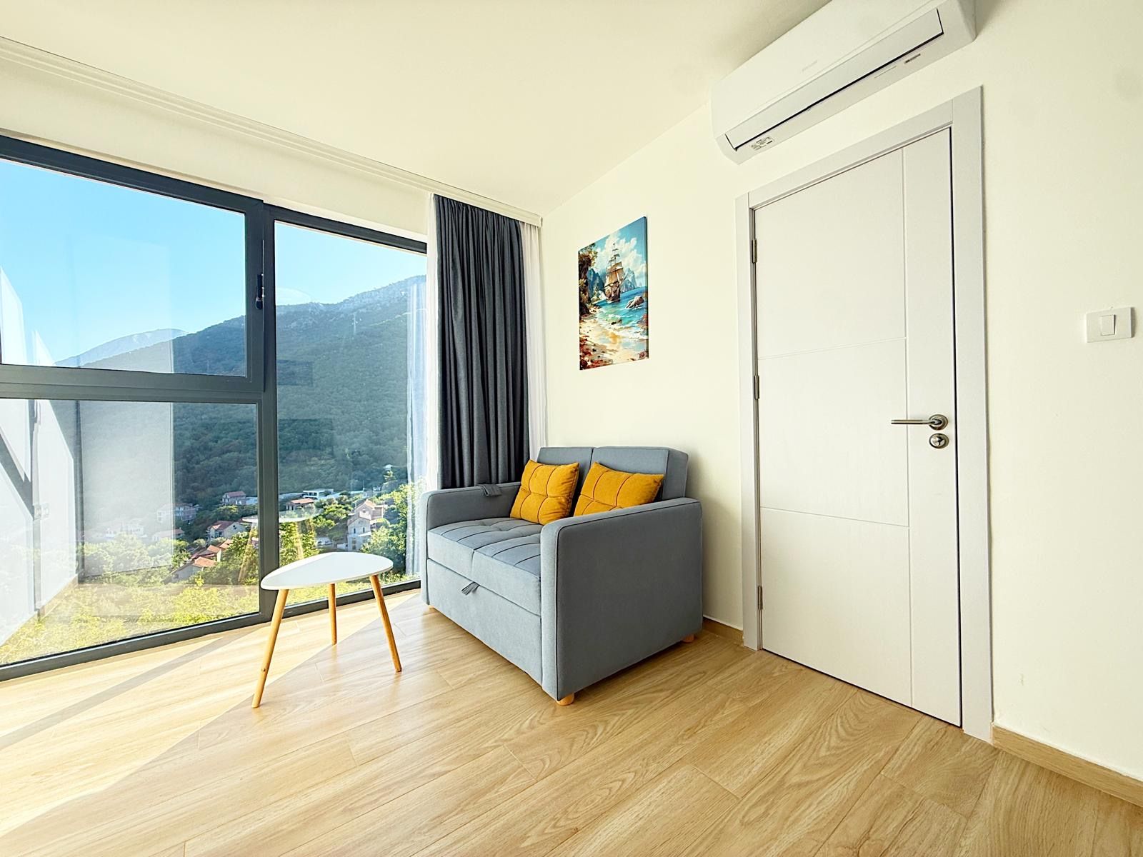 Ein modernes Zimmer mit einer grauen Couch, einem kleinen weißen Tisch und einem großen Fenster mit Blick auf eine gebirgige Landschaft.