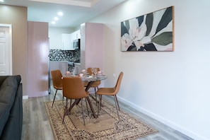A dining area with a table set for four, surrounded by chairs, and a kitchen in the background. - Gulf Residence Apt2 (La Romain)