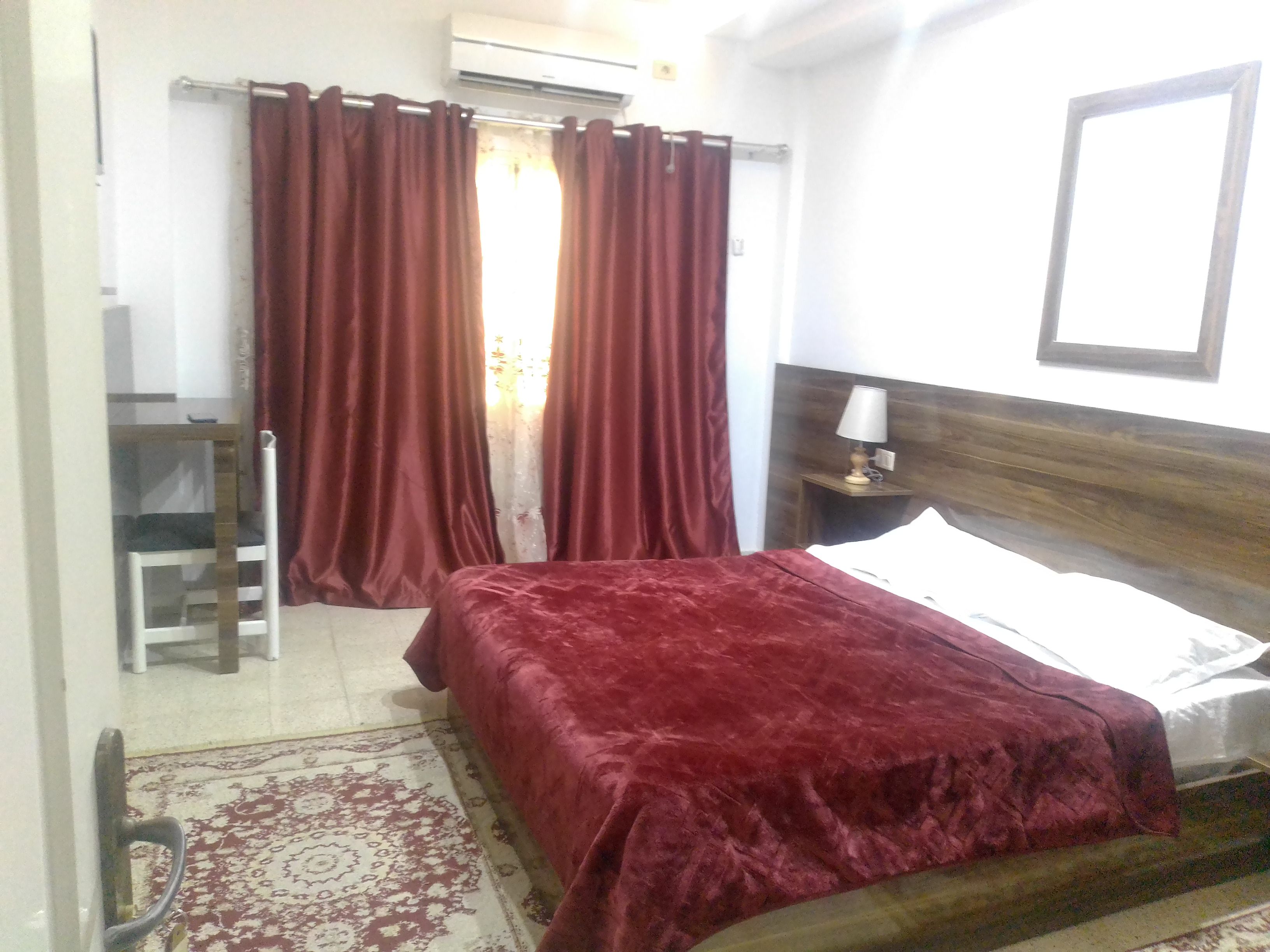 A bedroom with a bed, a nightstand, a lamp, a chair, a desk, and a window with curtains.