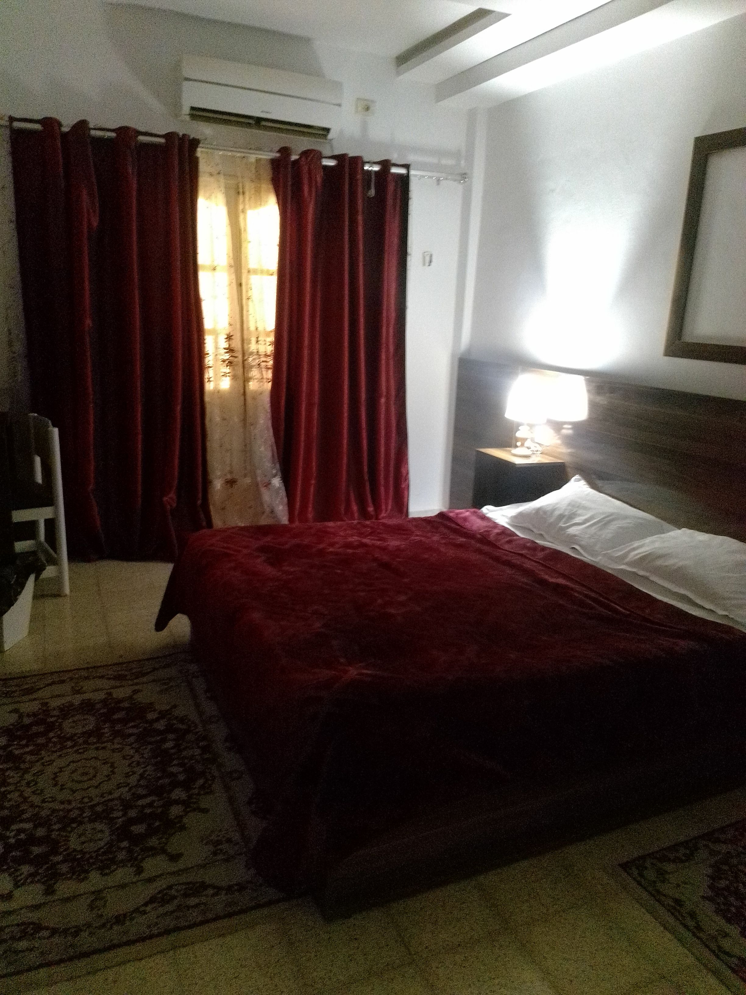 A hotel room with a bed, a nightstand with a lamp, a window with curtains, and a door.