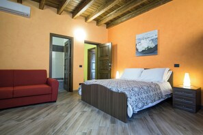 A bedroom with a wooden ceiling, a bed with patterned bedding, a red sofa, and a bedside table with a lamp. - New Listing (Malvagna)