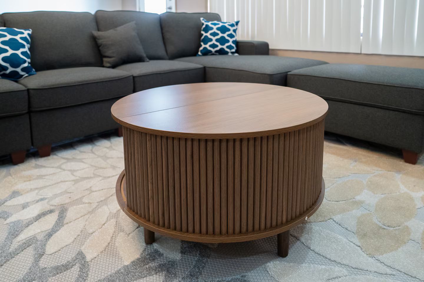 A round wooden side table with vertical slats, placed on a patterned carpet in a living room with a grey sectional sofa and patterned cushions.