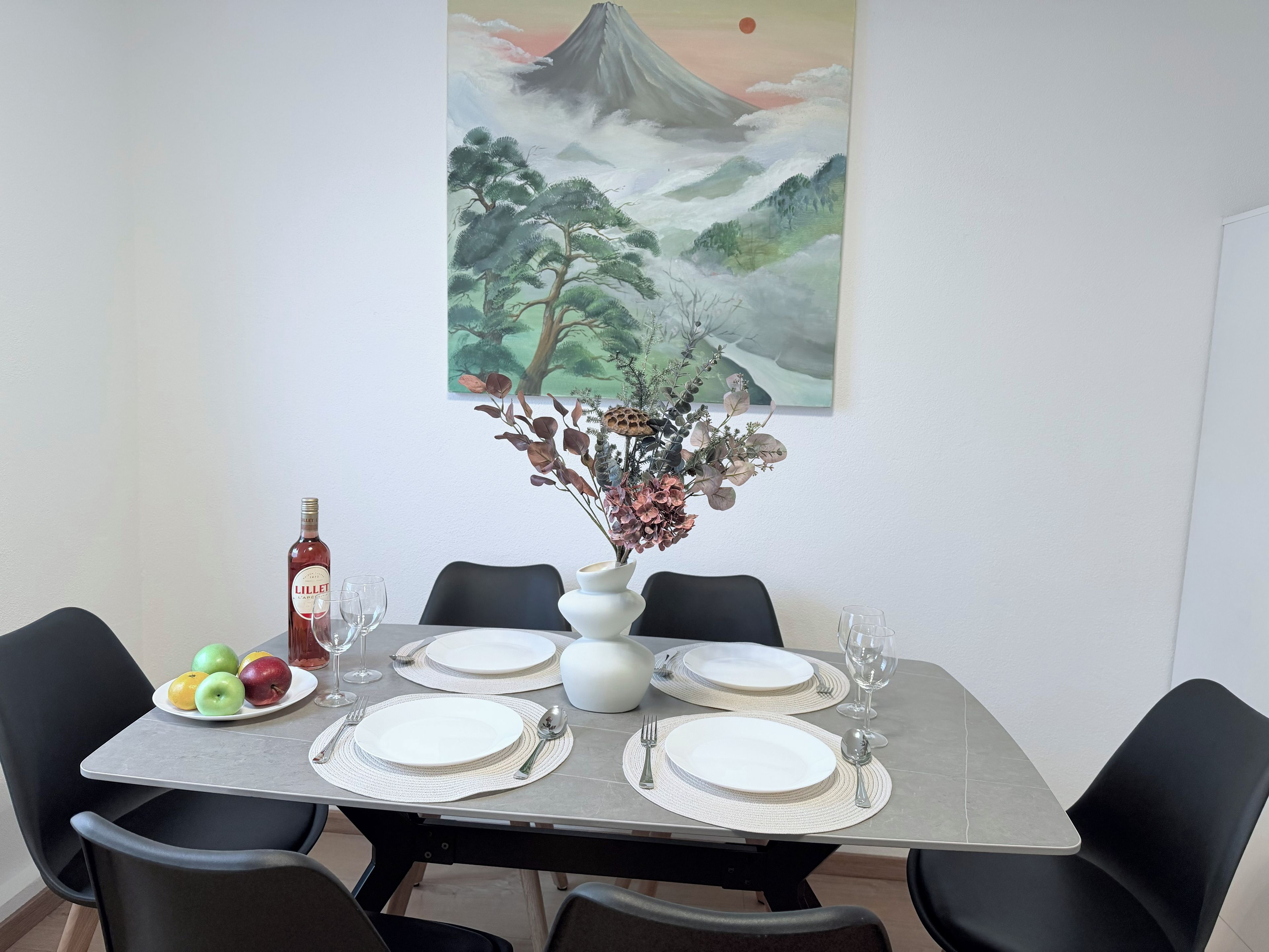 A dining table set with plates, cutlery, and a bottle of liquor, with a painting of a mountain landscape above.