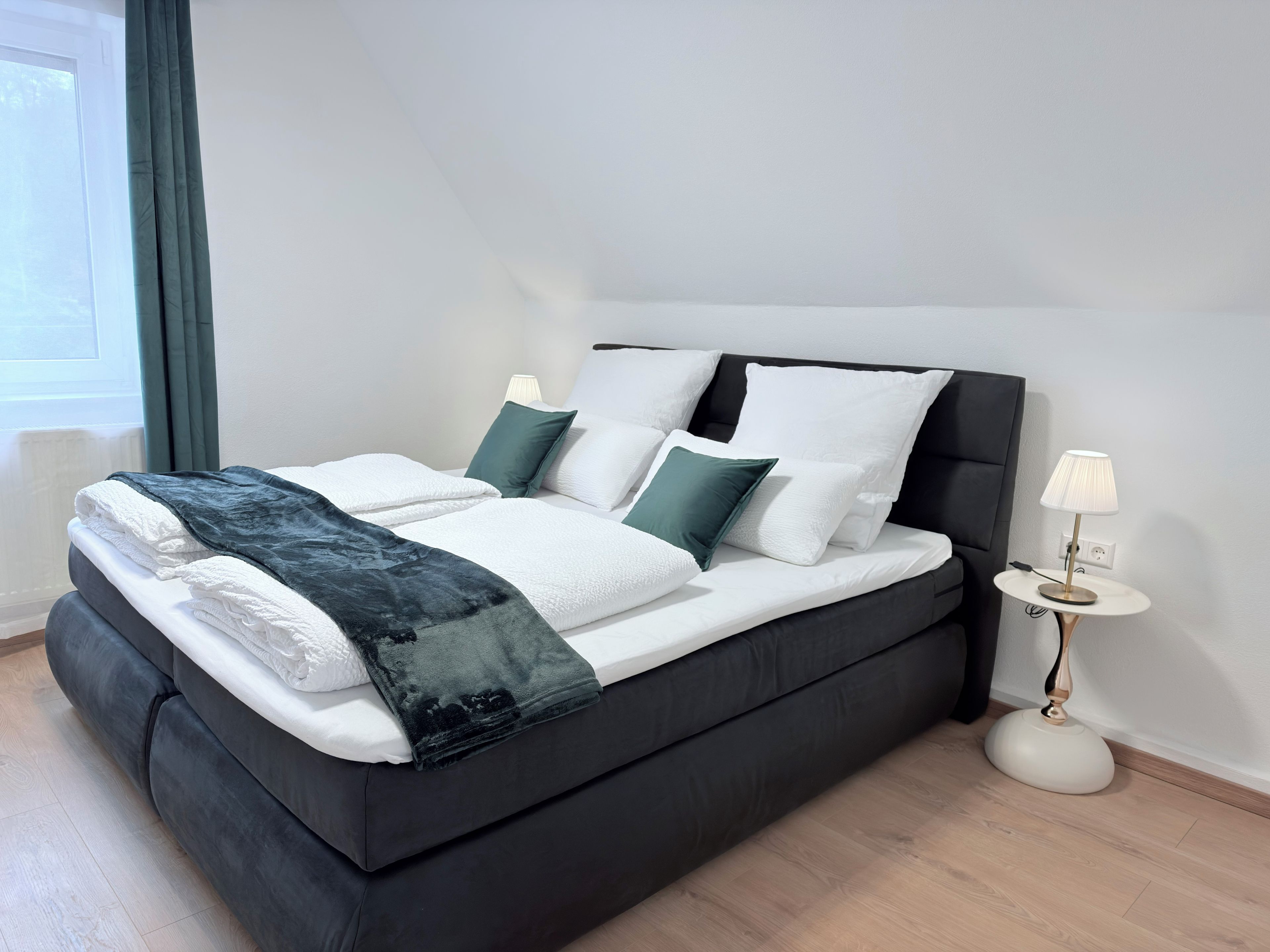 A modern bed with a dark headboard, white bedding, and a mix of green and white pillows. A bedside lamp is on a small table beside the bed.