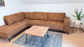 A sectional sofa with a matching ottoman, a small wooden coffee table, a patterned rug, and a potted plant on a wooden floor.