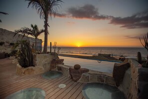 A sunset view from a wooden deck with a pool, palm trees, and a stone wall.