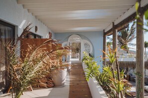 A covered outdoor corridor with potted plants, a wooden structure, and a view into a room with a white ceiling.