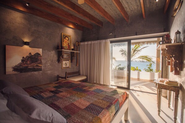 A bedroom with a large bed, a nightstand, a window with curtains, and a view of the sea.
