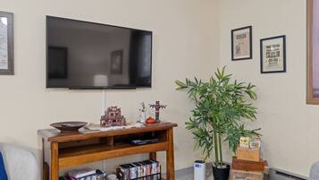 Un soggiorno con una TV a schermo piatto montata a parete, un mobile in legno contenente libri e oggetti decorativi, una grande pianta in vaso e certificati incorniciati appesi al muro.