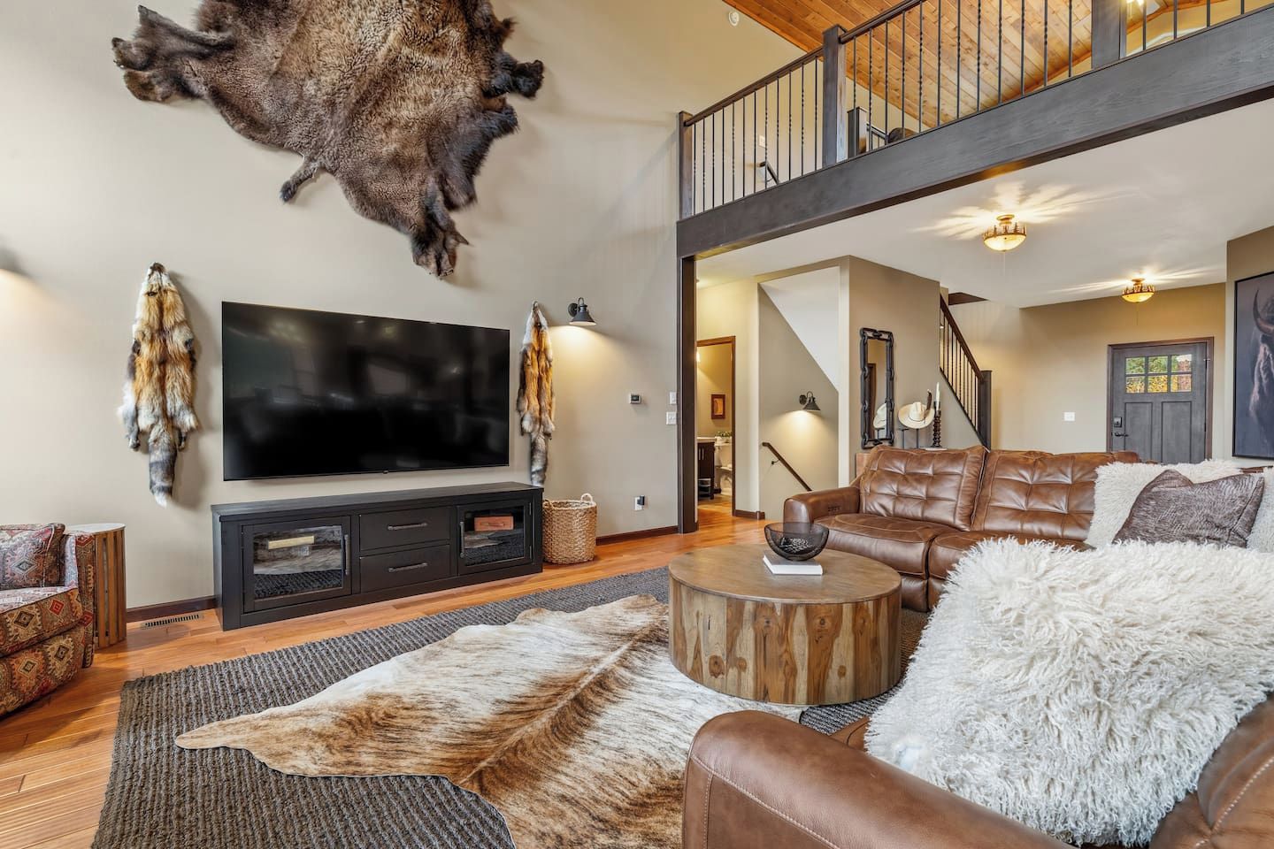 A spacious living room with a large flat-screen TV, leather sofas, a round wooden coffee table, and a mounted animal hide on the wall.