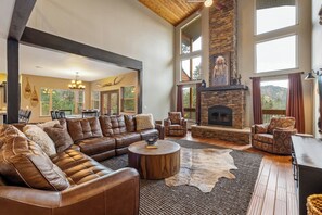 A spacious living room with a leather sectional sofa, a stone fireplace, and a large window overlooking a scenic view. - Private: 10 Beds, Hot Tub & Forest Views Near Lead (Lead)