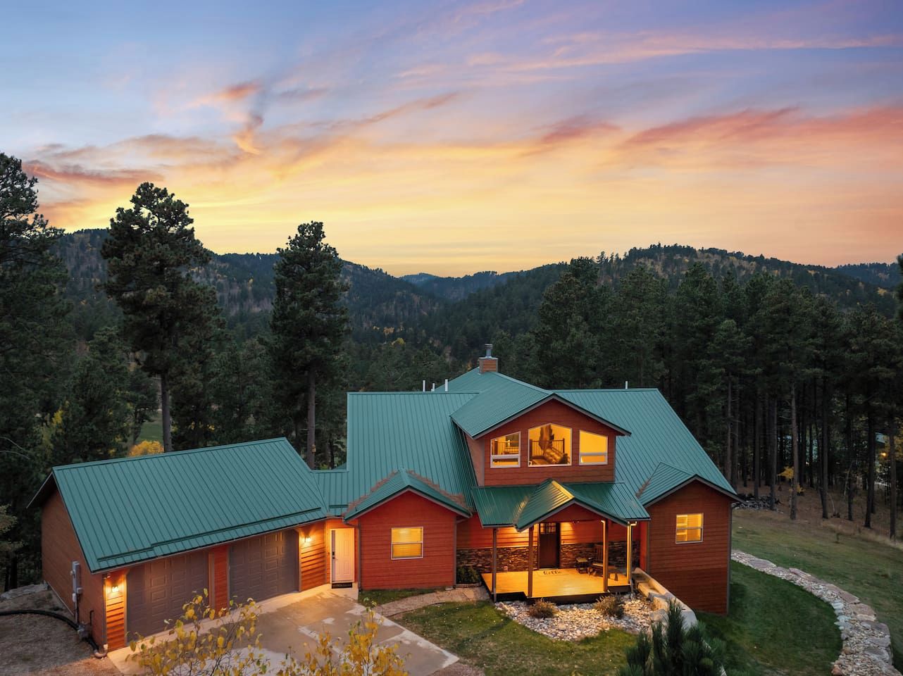 A two-story log cabin with a green metal roof, a covered front porch, and a stone fireplace.