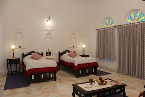 Une chambre avec deux lits en bois, chacun recouvert d’une couverture rouge et de draps blancs à motifs de dentelle. On y trouve deux tables de chevet avec des lampes, une petite table avec une théière et des fruits, et des rideaux à motifs.