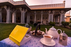 Une table dressée dans une cour, avec un livre, une théière, une tasse et des biscuits, et un bâtiment en arrière-plan.