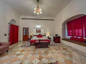 Ein geräumiges Schlafzimmer mit einem großen Bett, einem Deckenventilator, einer Chandelierleuchtre und einem Fenster mit roten Vorhängen.
