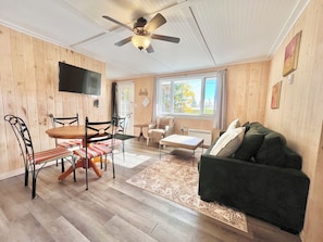 Un salon chaleureux avec un plancher en bois, un canapé vert, une table à manger avec des chaises, un ventilateur de plafond et un téléviseur fixé au mur.