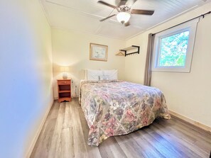Une chambre à coucher comprenant un lit, une table de chevet, un ventilateur de plafond, une fenêtre et une étagère fixée au mur.