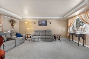 Una sala de estar con un sofá gris, un sillón más pequeño, una mesita de centro y una mesita auxiliar. Hay un cuadro en la pared y una lámpara de pie.