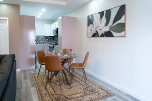 A dining area with a table set for four, surrounded by chairs, and a kitchen in the background. - Gulf Residence Apt5 (La Romain)