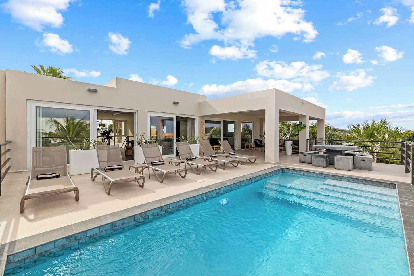 Ett område vid poolen med solstolar, en pool och en möblerad sittgrupp under tak med ett bord och stolar.