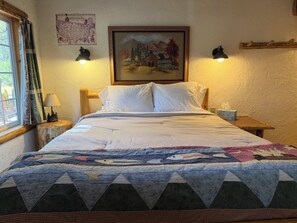 Ein Bett mit einer Stegdecke, ein Nachttisch mit einer Lampe, ein Gemälde an der Wand und ein Fenster mit Vorhängen.