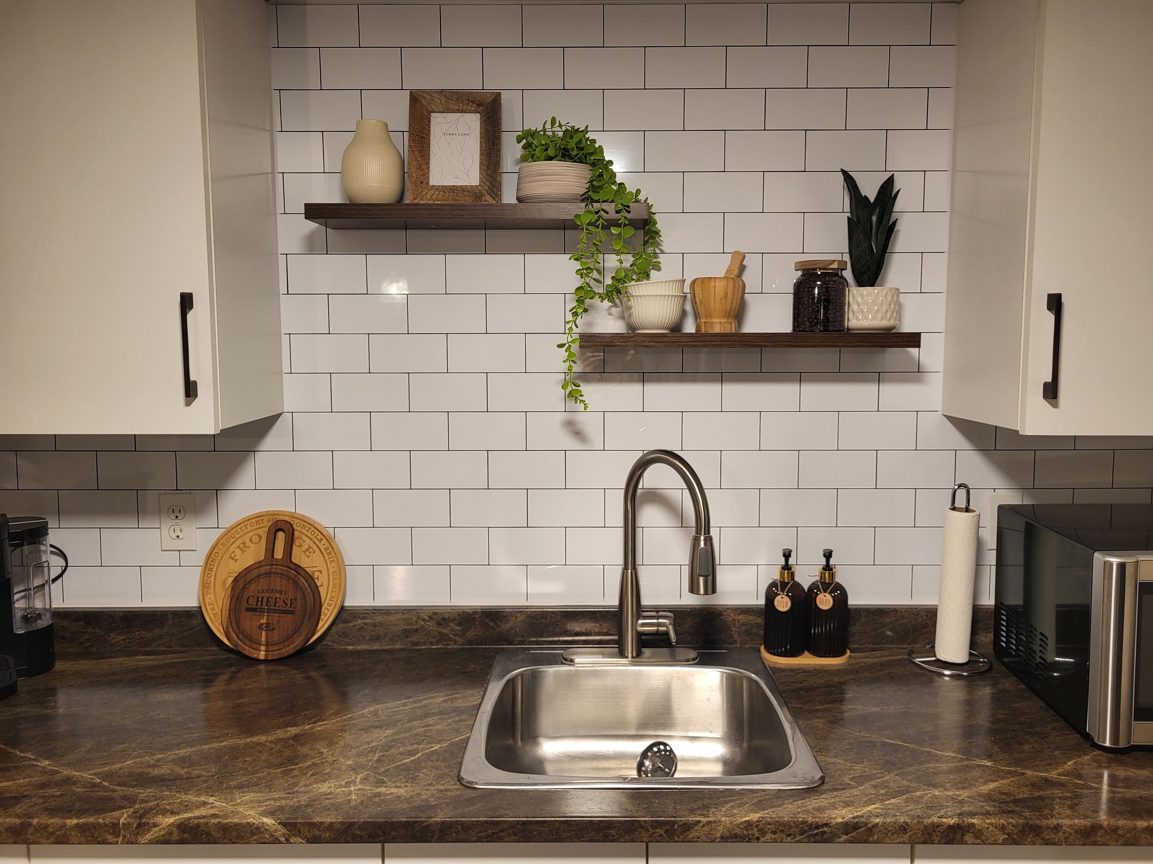 A kitchen with a stainless steel sink, white tiled backsplash, and wooden shelves with decorative items.