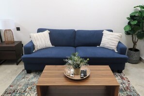 En blå sofa med to hvide puder, et træsofabord med en plante og fjernbetjeninger, en lampe og en potteplante.