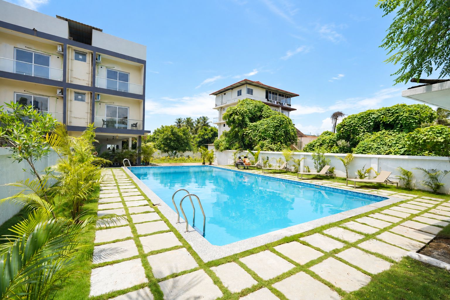 A swimming pool with a tiled deck, surrounded by greenery and a white wall.