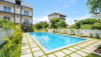 A swimming pool with a tiled deck, surrounded by greenery and a white wall.